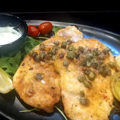 Plated dish of fried chicken with capers on a bed of greens with lemon wedges and tomatoes.