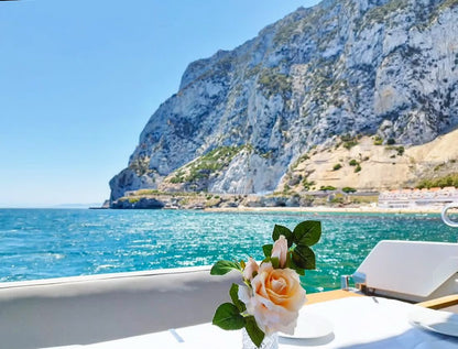 Table with a flower arrangement on a yacht against a scenic backdrop of blue water and rocky cliffs of Gibraltar.