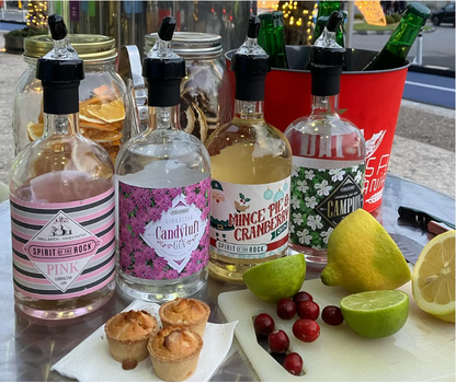 Bottles of gin with festive labels, mince pies, and fruit on a table outdoors.