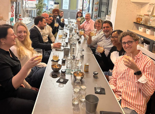 Group of people sitting at a long bar counter, raising glasses in a toast, on a gin tasting tour.